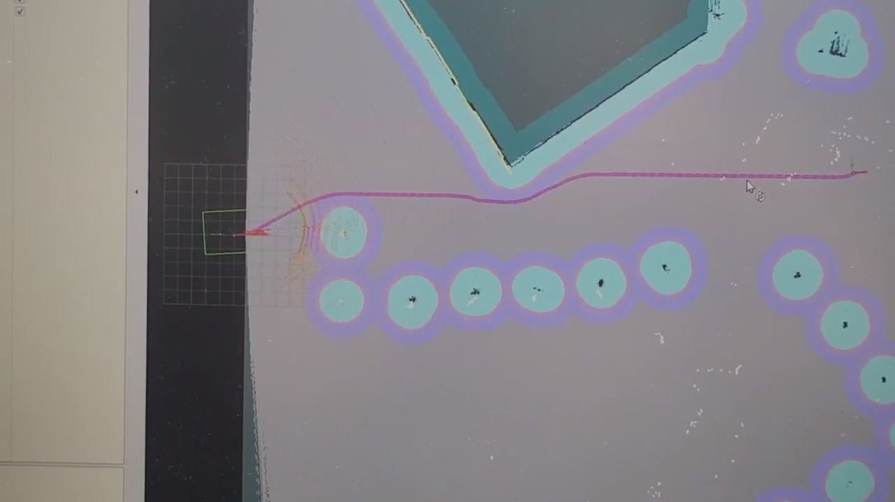 Photograph of a monitor showing an RViz navigation view with a grey global costmap, cyan inflation circles around obstacles, a dark blue mapped wall, and a magenta planned path running from the robot footprint to a goal on the right