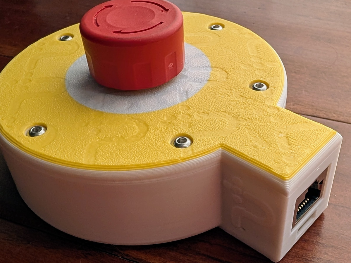Photograph of the assembled Protective Stop remote: a yellow and white 3D printed enclosure with a large red twist-release mushroom button on top, and an RJ45 Ethernet jack visible in the side pod