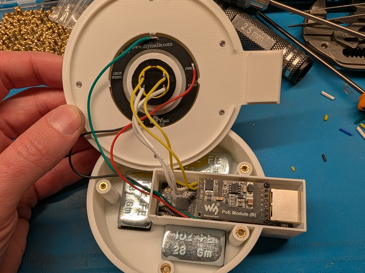 Photograph of the open enclosure with the lid folded back: a black Waveshare PoE Module (B) is screwed onto the ESP32-S3-ETH board in the side pod, and red, black, green, white and yellow wires run from the board up to the LED ring and E-stop button in the lid