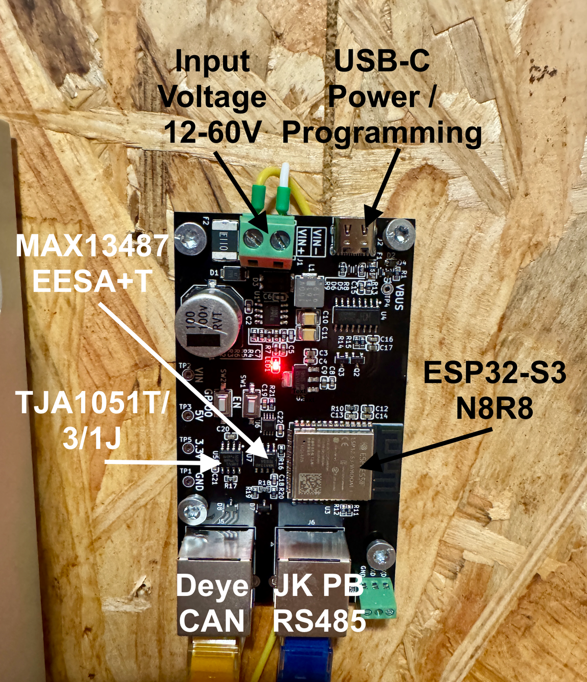 Photo of a custom black ESP32-S3 PCB screwed to a plywood wall with annotations naming the input voltage terminal, USB-C port, MAX13487 and TJA1051T transceivers, the ESP32-S3 module, and two RJ45 jacks labelled Deye CAN and JK PB RS485