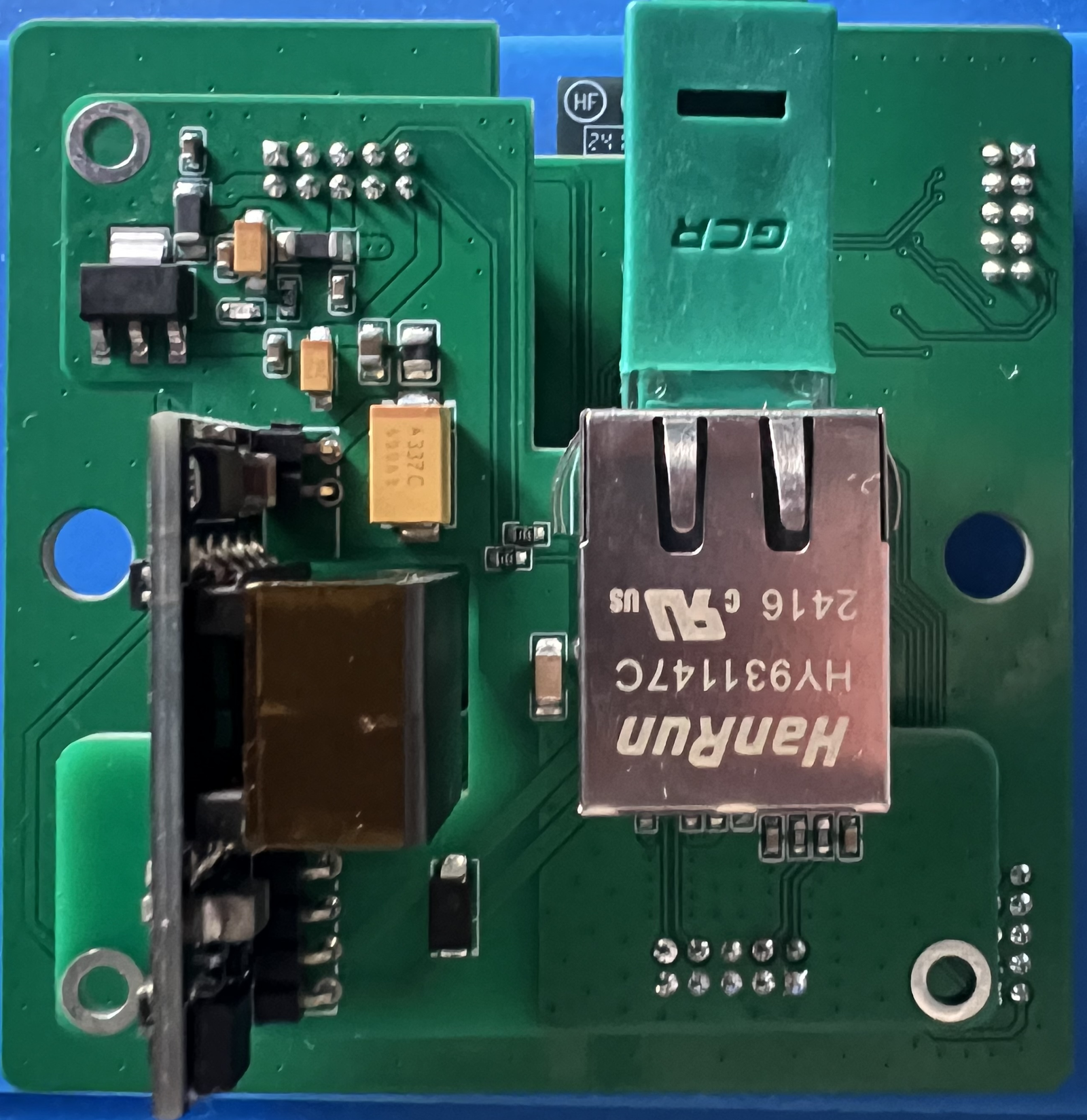 Photograph of the back board showing the metal HanRun HY931147C RJ45 jack with a green Ethernet plug inserted, the black SDAPO PoE module with its copper-taped transformer, and an AMS1117 regulator