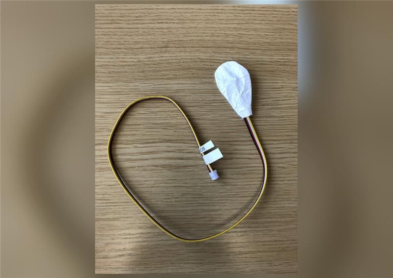 A temperature and humidity probe photographed on a wooden desk, with a coiled four-conductor ribbon cable, a small white connector, a label tag and a white Tyvek sleeve covering the sensor head