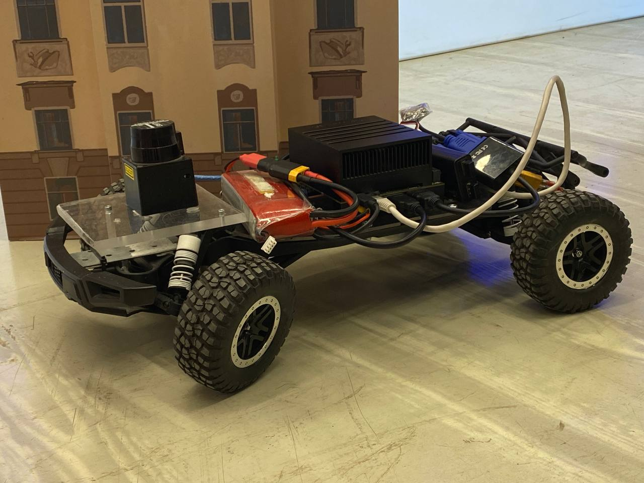 Photograph of the ULSTU buggy robot, a black short-course radio-controlled car chassis with a scanning laser on an acrylic plate at the front, a red lithium battery, a black finned computer, and a grey Ethernet cable looping over the rear, standing on a laboratory floor in front of a cardboard building model