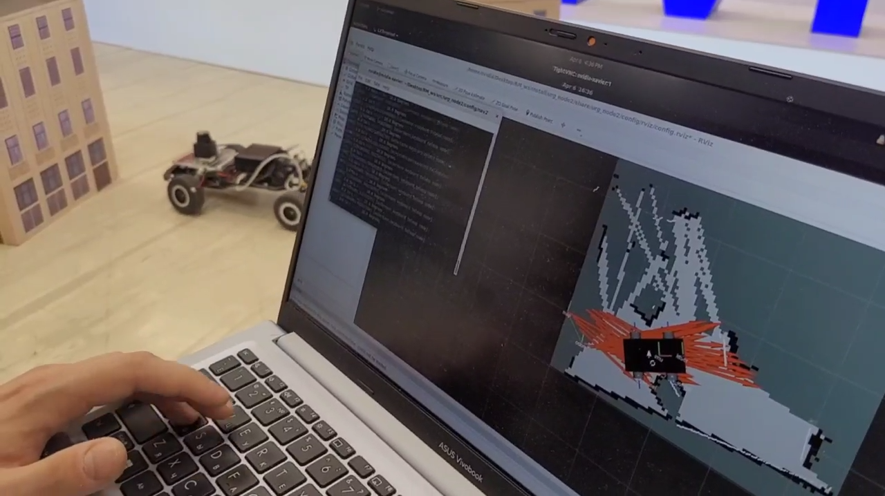 Photograph of a laptop screen showing an RViz window with a partly built black and white occupancy map and red odometry arrows, with the buggy robot and cardboard building models visible on the floor behind the laptop