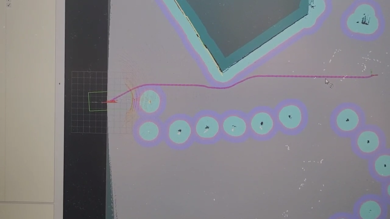 Photograph of a monitor showing an RViz navigation view with a grey global costmap, cyan inflation circles around obstacles, a dark blue mapped wall, and a magenta planned path running from the robot footprint to a goal on the right