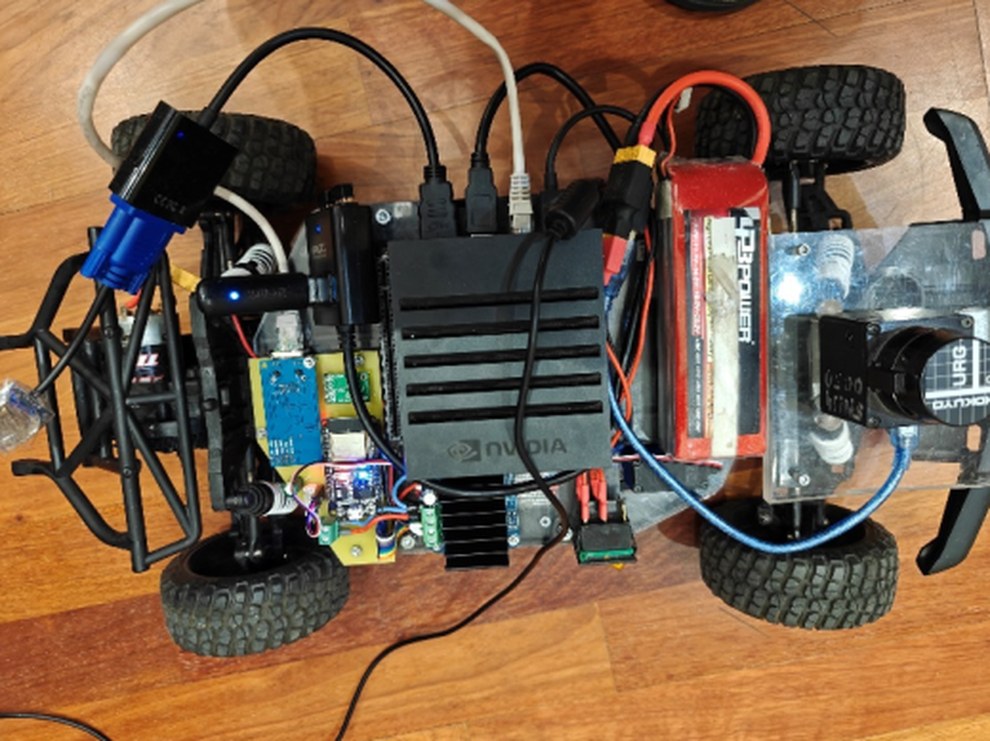 Top-down photograph of the robot on a wooden floor, showing the Nvidia onboard computer with its finned case in the centre, a red lithium battery beside it, the custom control board with its blue Ethernet module at the left, and the Hokuyo scanning laser on an acrylic plate at the right