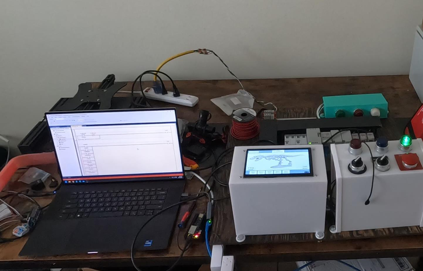 Photo of the Group 21 test bench: a laptop showing PLC ladder logic on the left, a white HMI enclosure displaying a T-Rex skeleton in the center, and a control box with indicator lights and an emergency stop on the right, with DIN rail hardware behind them
