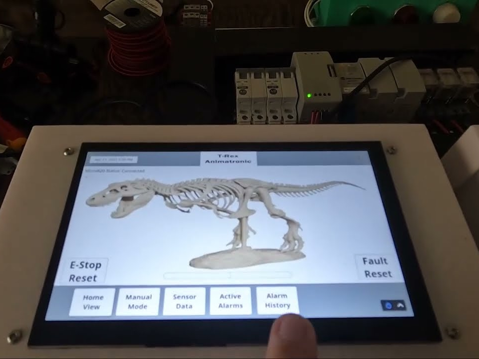 Close-up of a finger tapping the touch HMI, which shows a T-Rex skeleton, E-Stop Reset and Fault Reset buttons, and a status line reading Micro820 Status Connected, with the PLC DIN rail visible behind the panel