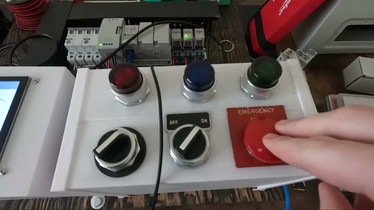 Close-up of the white control box with red, blue, and green indicator lights, two rotary selector switches, and a large red emergency stop button, with Allen-Bradley DIN rail modules in the background