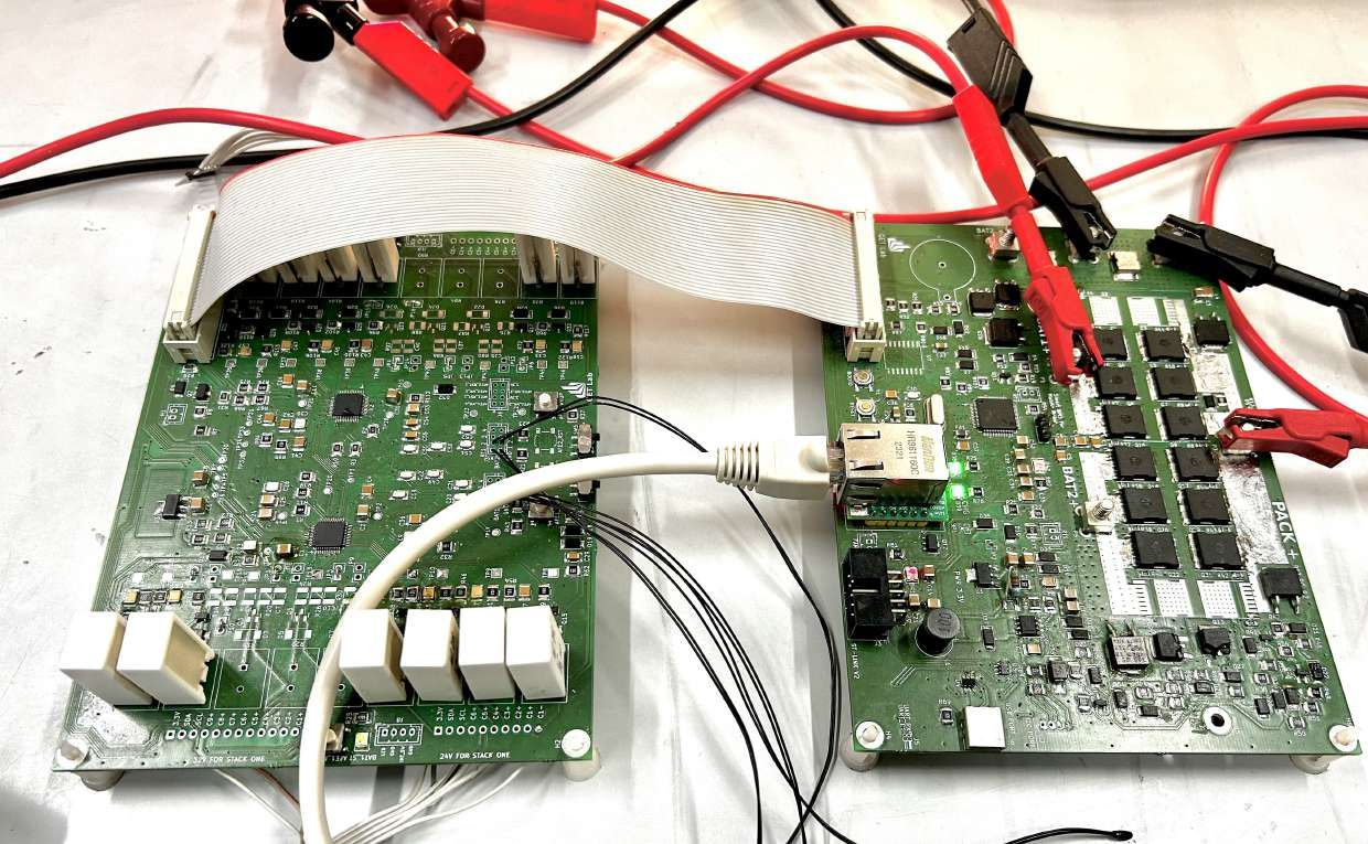 Bench photo of the two BMS boards joined by a grey ribbon cable, with red and black test leads clipped to the pack terminals and an Ethernet cable plugged into the RJ45 jack, its green link LED lit