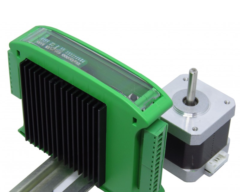The green GEVINO Motor enclosure on a DIN rail next to a NEMA-frame stepper motor, showing the transparent front window and the side terminal blocks